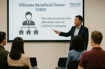 A professional seminar hosted by Thai-Co Consultants, where a speaker is presenting a slide explaining “Ultimate Beneficial Owner (UBO)” to a seated audience. The presenter points to a projected diagram showing a person controlling multiple entities, emphasizing the importance of UBO transparency.